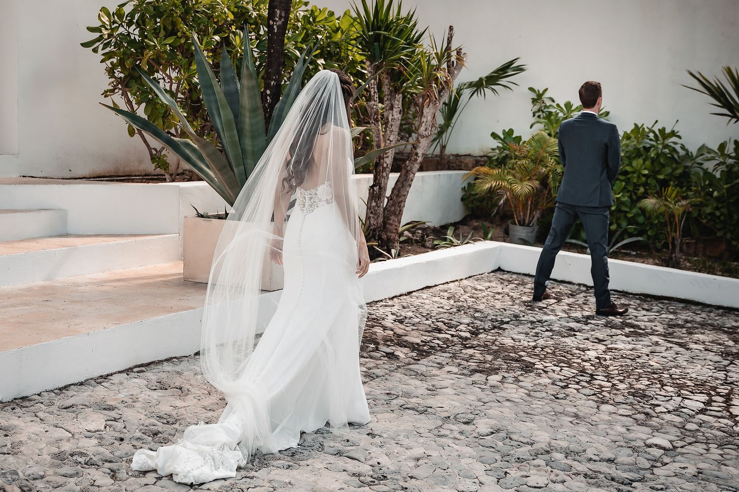 Bride in white gown and veil stands near steps as groom in blue suit walks ahead at outdoor wedding venue.