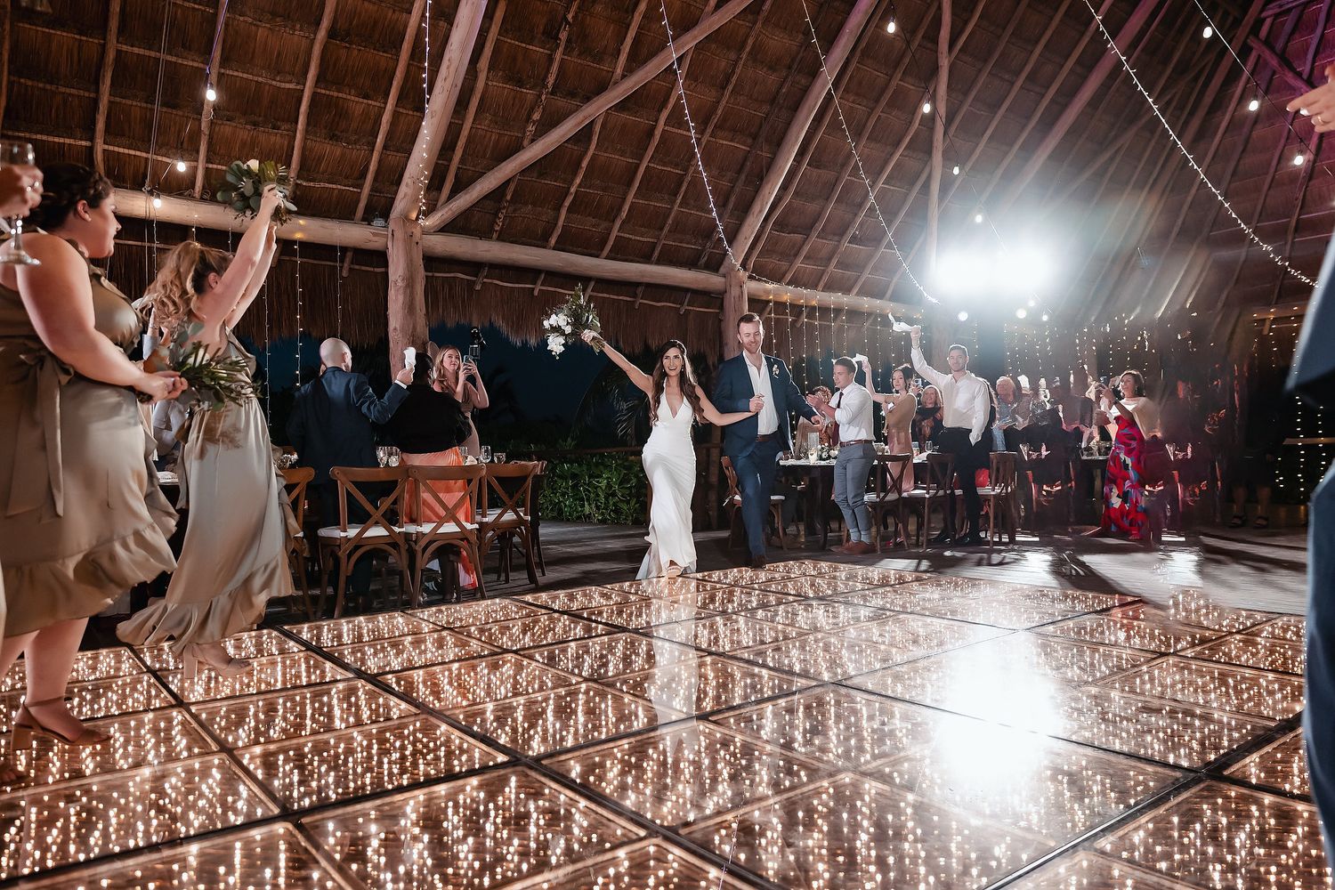 Bride and groom enter reception on a sparkling mirror dance floor surrounded by guests cheering under a warmly lit outdoor venue.