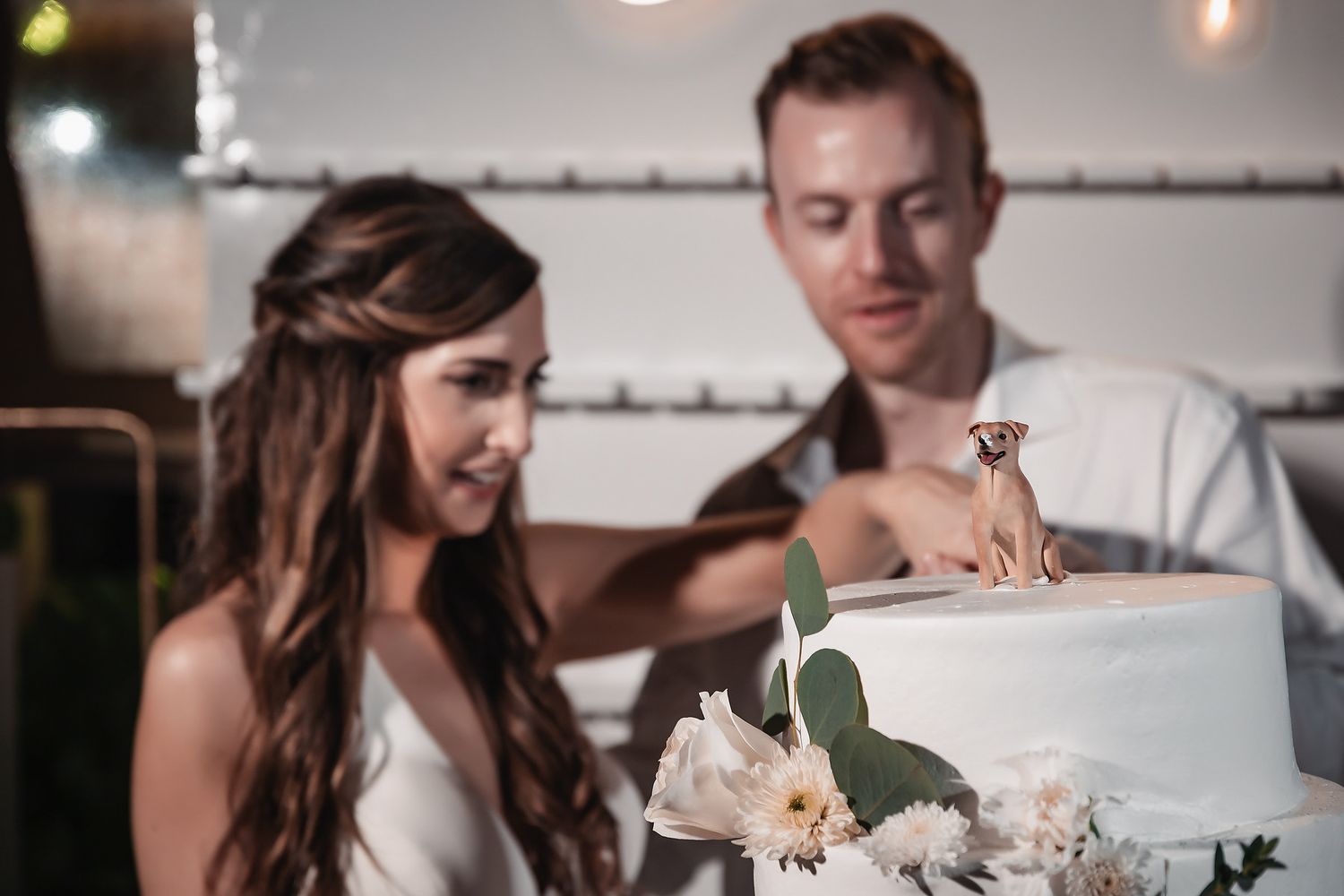 Bride and groom cutting their white wedding cake decorated with flowers and a dog topper.