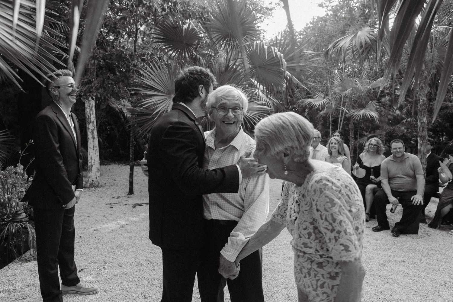 Guests greeting each other at an outdoor wedding ceremony surrounded by tropical plants.