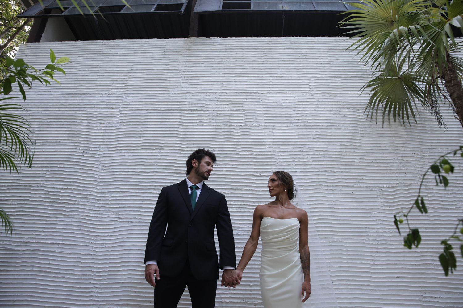 Bride and groom holding hands in front of a white textured wall surrounded by tropical palms on their wedding day.