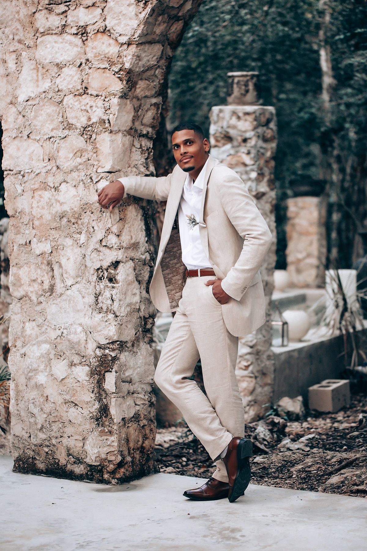 Stylish groom in cream suit poses against stone ruins in outdoor setting.