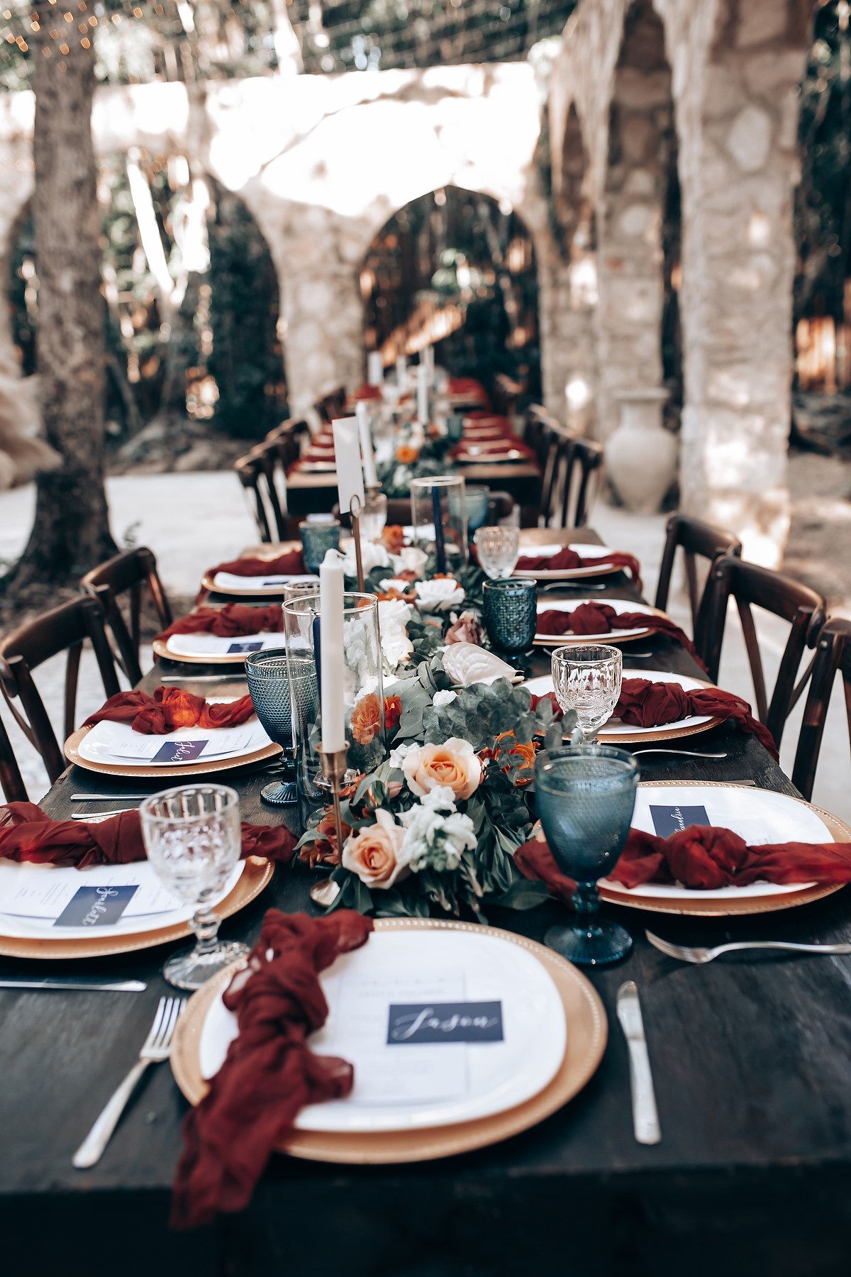 Formal table setting with burgundy napkins, gold chargers, and floral centerpieces on black tablecloths.