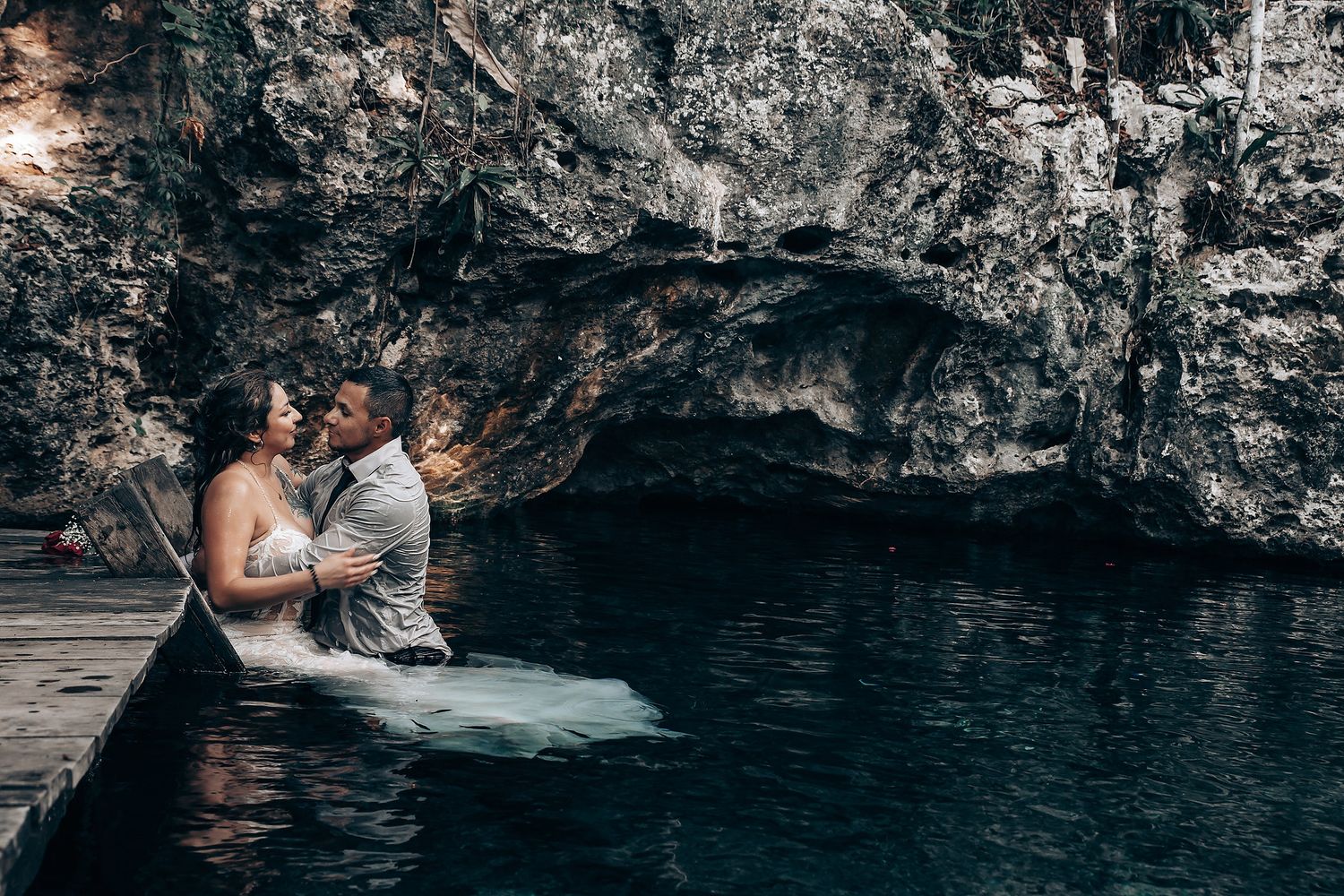 A couple sits together at the edge of a serene cenote cave pool with dark rocky walls in Mexico.