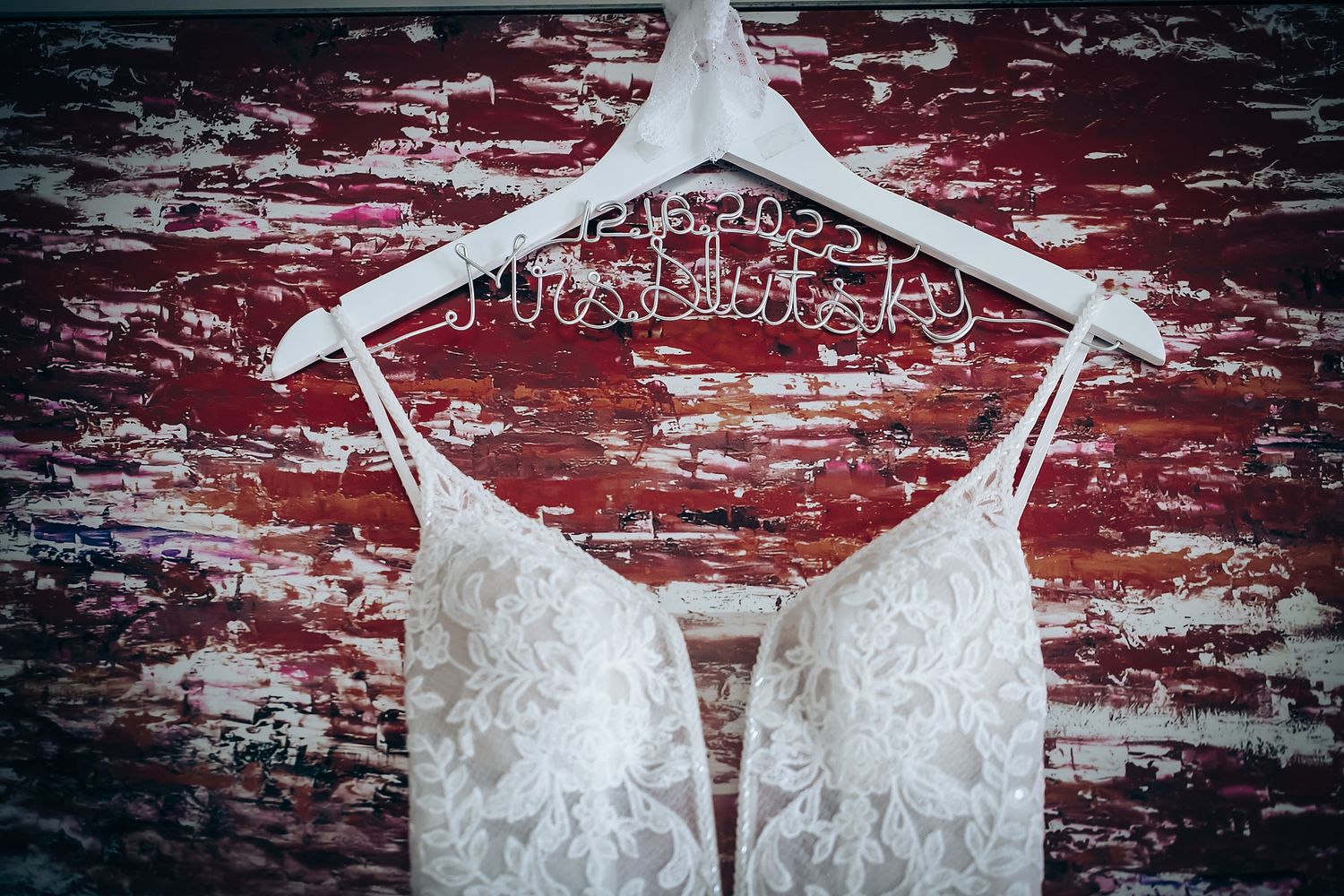 Elegant white lace wedding dress hanging on personalized hanger against rustic red background.