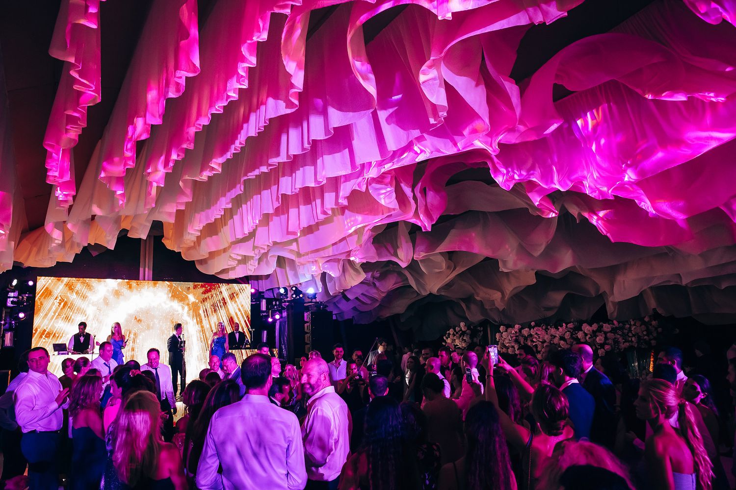 Wedding venue ceiling illuminated with vibrant pink lighting and white fabric draping.