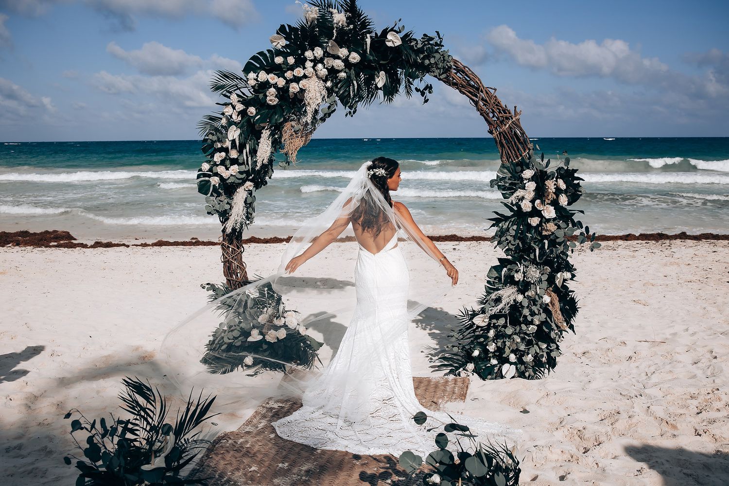 Romantic beach wedding ceremony setup with floral arch and white chairs against turquoise ocean waves and blue sky.
