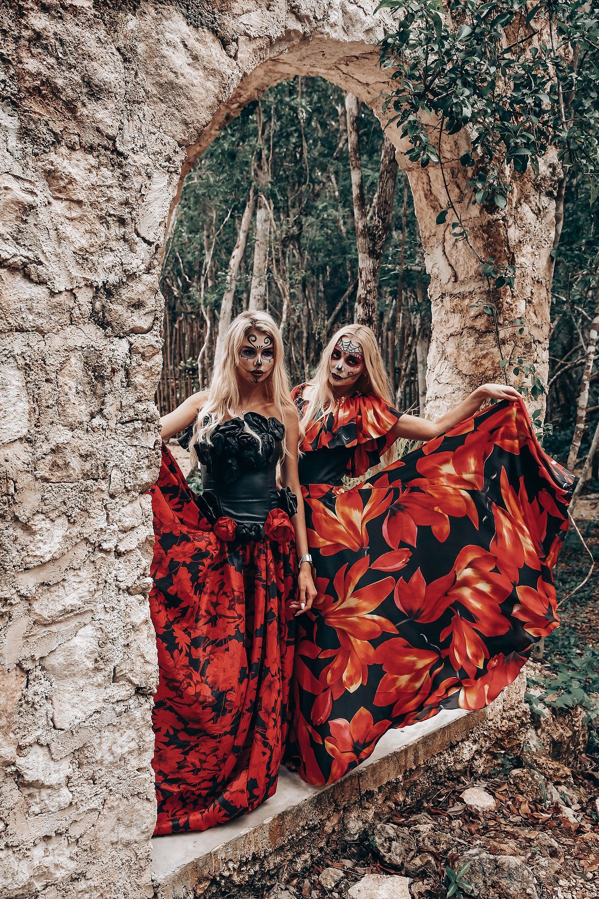 Two people in dramatic red and black dresses pose against stone archway in forest setting.