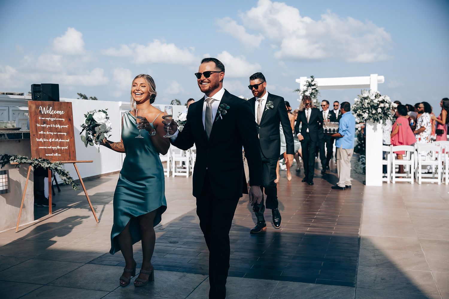 Wedding party members walk down an outdoor deck venue with white chairs and ocean views in the background.