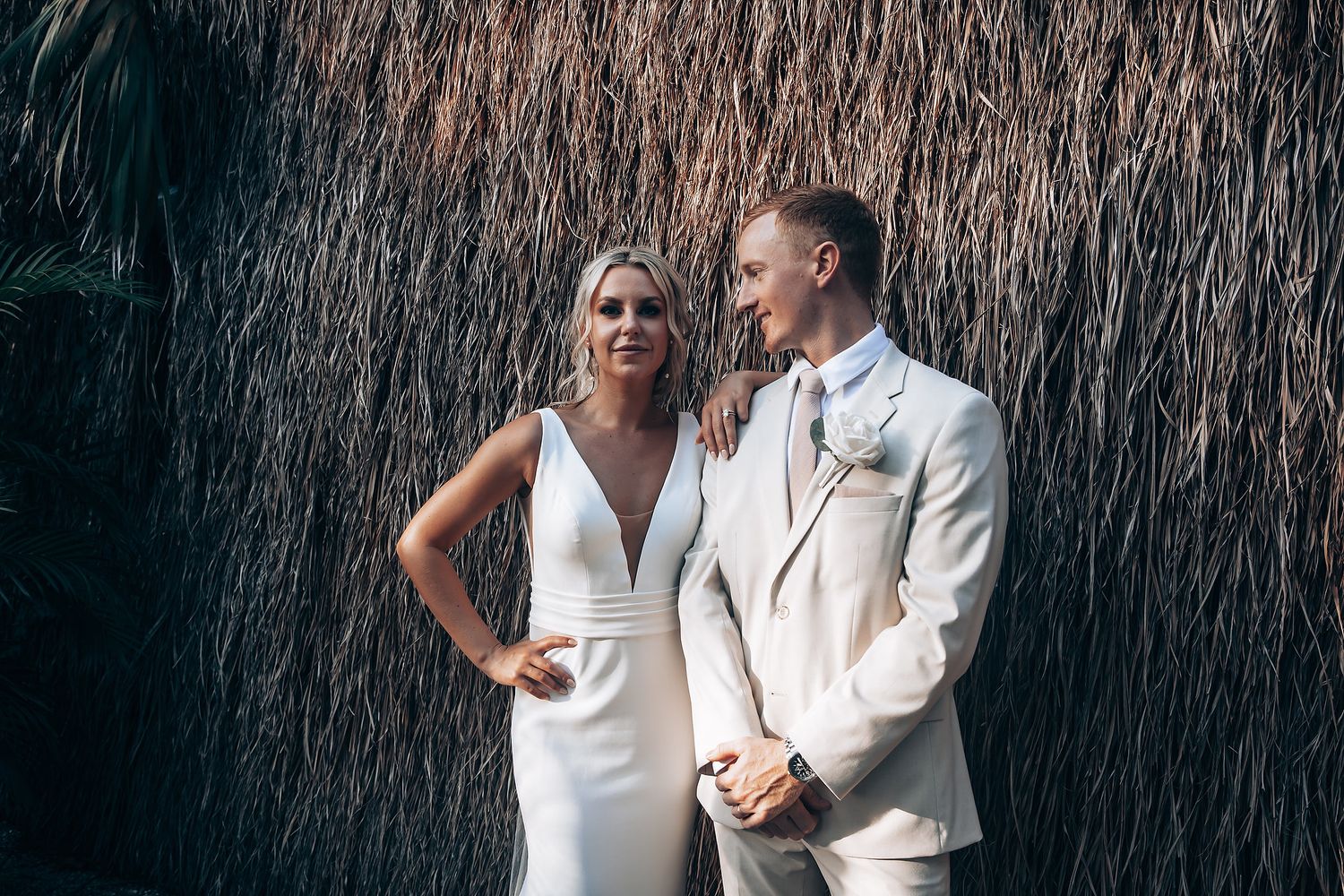 A couple in white attire poses intimately against a dark wooded backdrop during their wedding photoshoot.