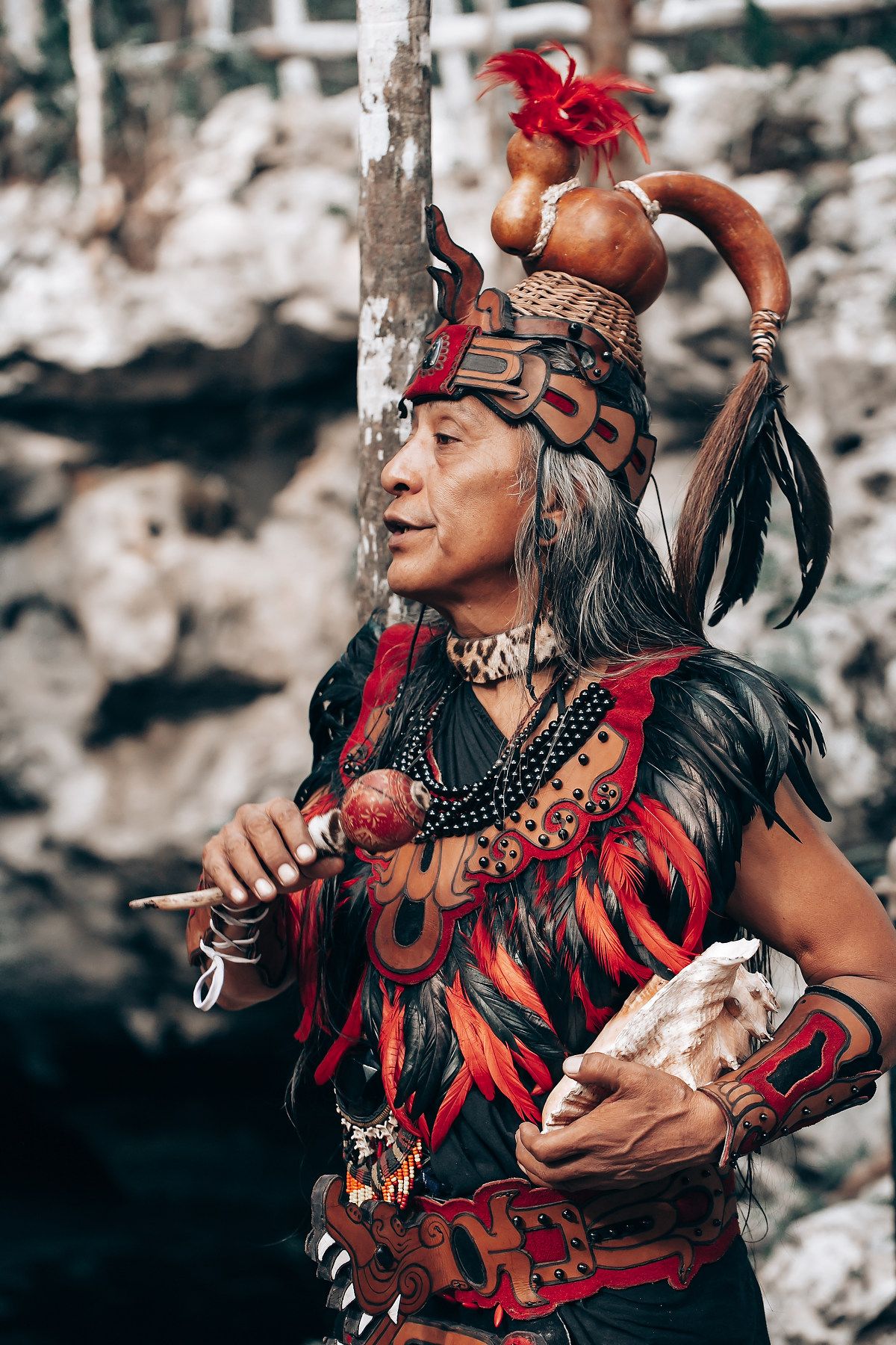 Traditional Hawaiian dancer performing in vibrant red feathered costume against rocky backdrop.