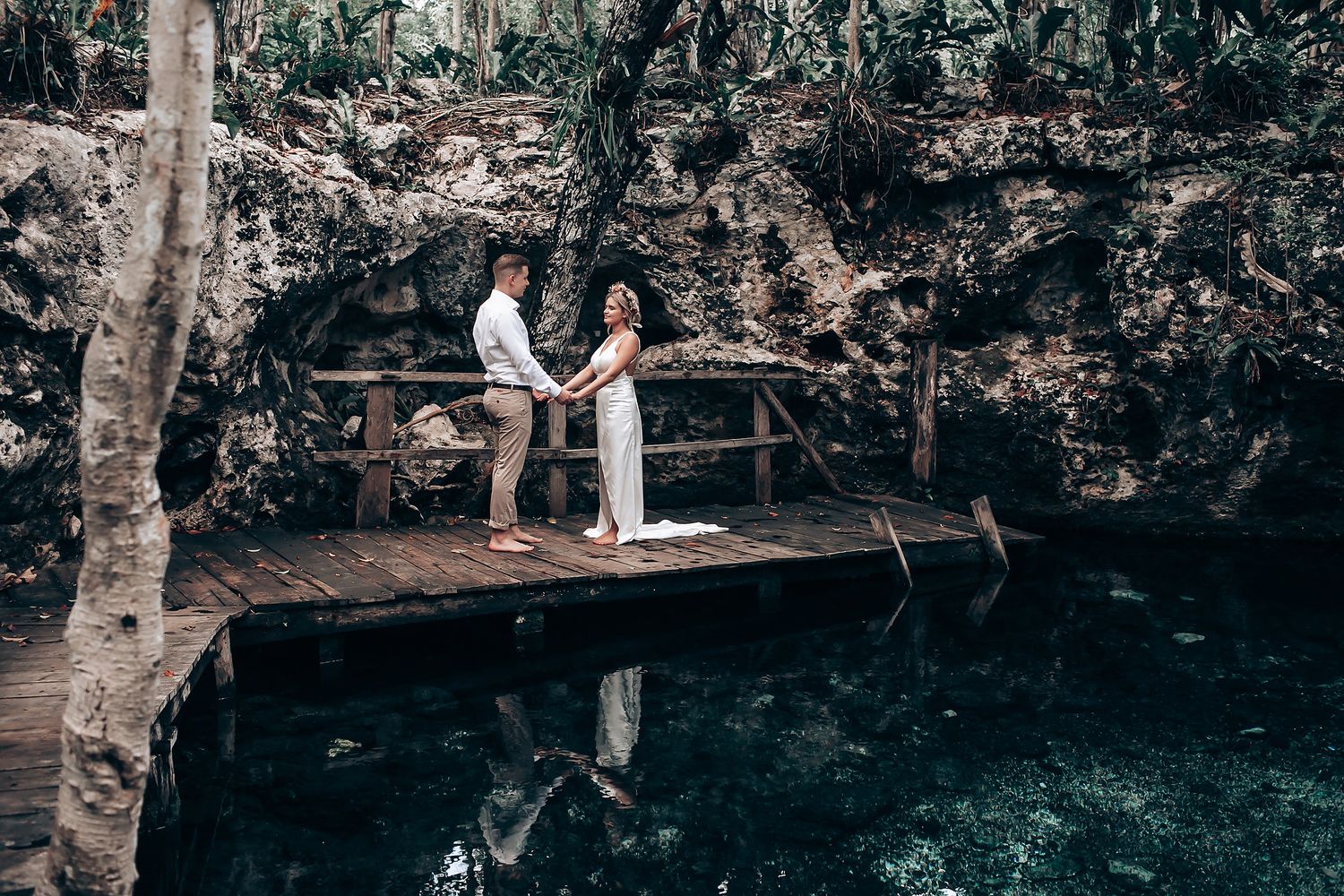 A couple shares an intimate moment on a wooden platform over a cenote surrounded by tropical vegetation.