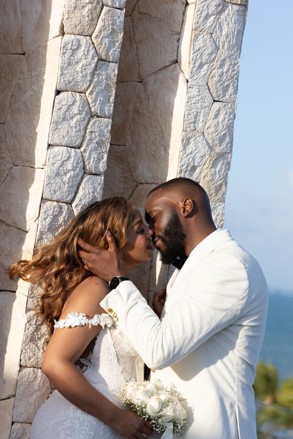 Bride and groom sharing a kiss beside white stone columns at seaside wedding venue.