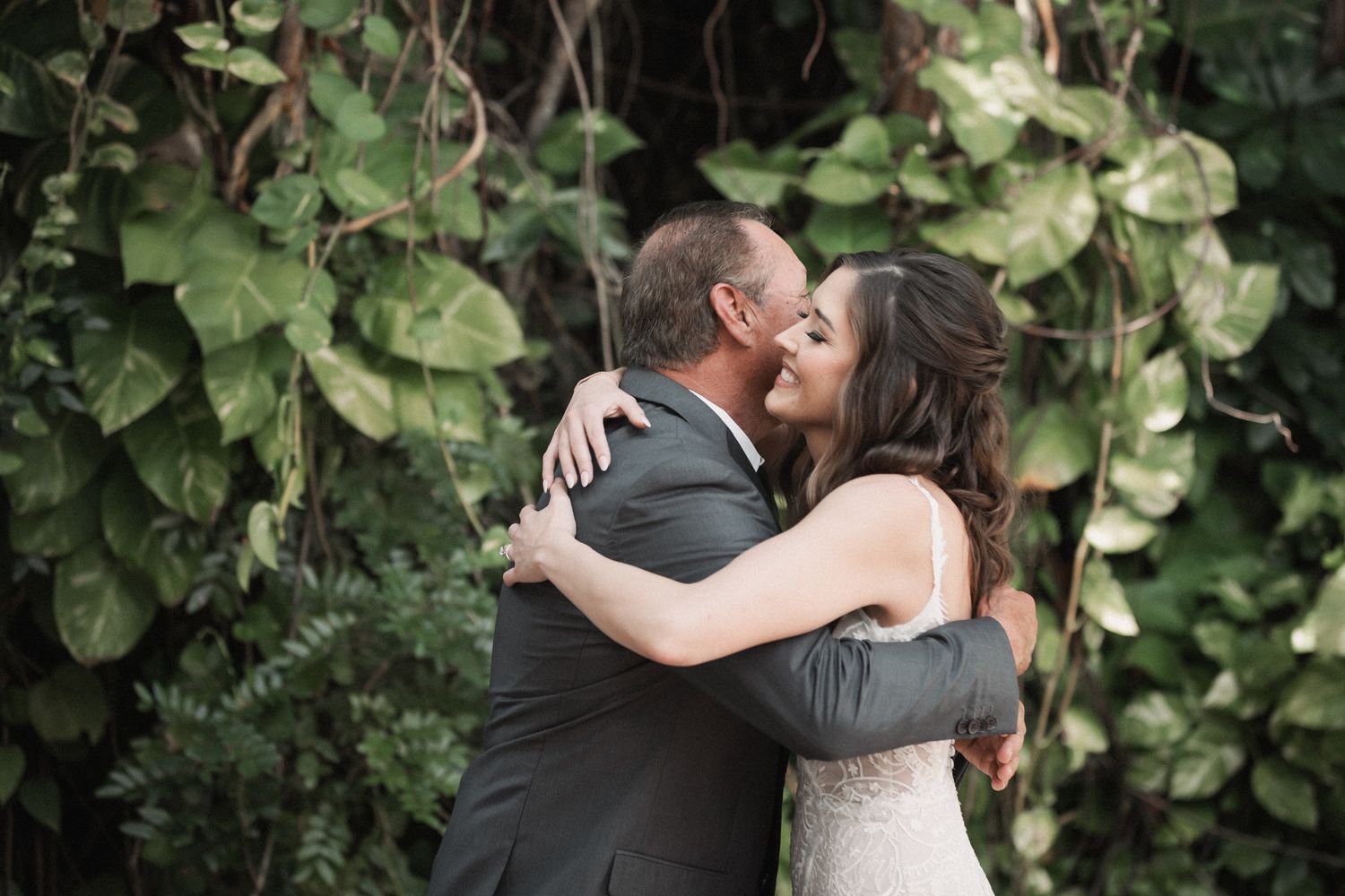 Bride sharing a joyful first look hug with her father surrounded by lush green vines.