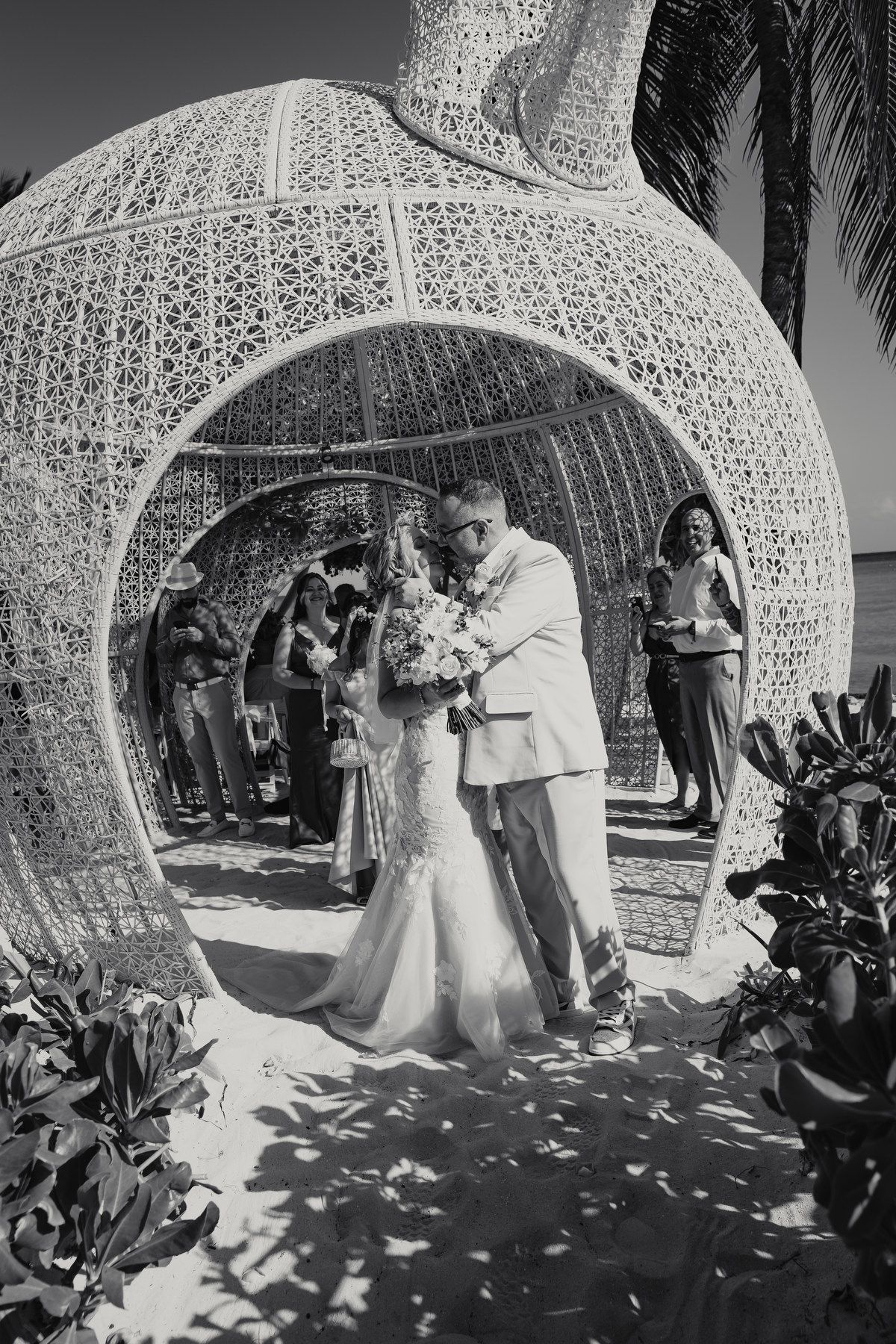Bride and groom share first kiss under ornate lattice arch on sandy beach wedding ceremony.