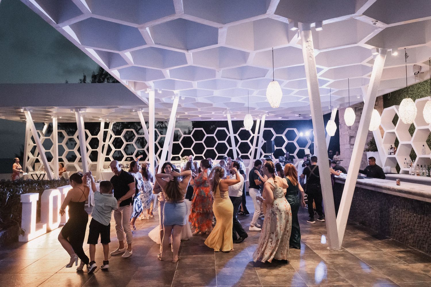 Wedding guests dance under geometric honeycomb ceiling at elegant outdoor rooftop reception venue.