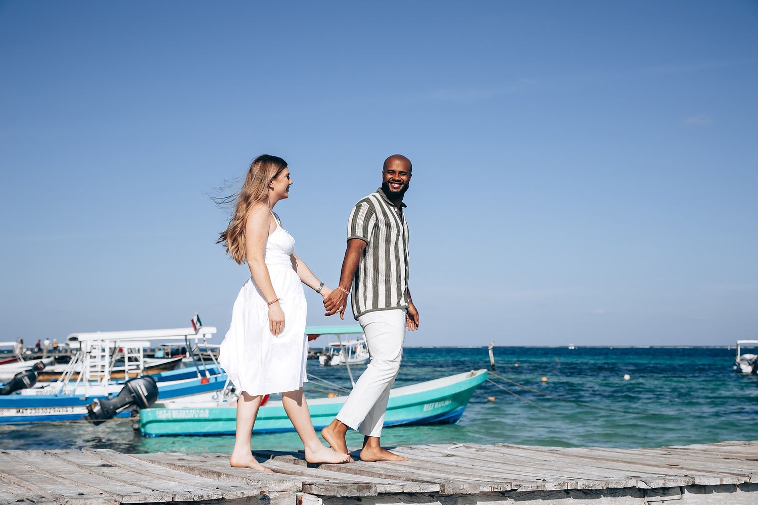 Interracial couple holding hands walking on a wooden pier by the ocean on a sunny day.