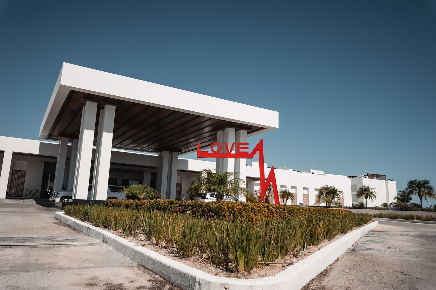 Modern building entrance with red sculptural sign, manicured shrubs, and clear blue sky in background.