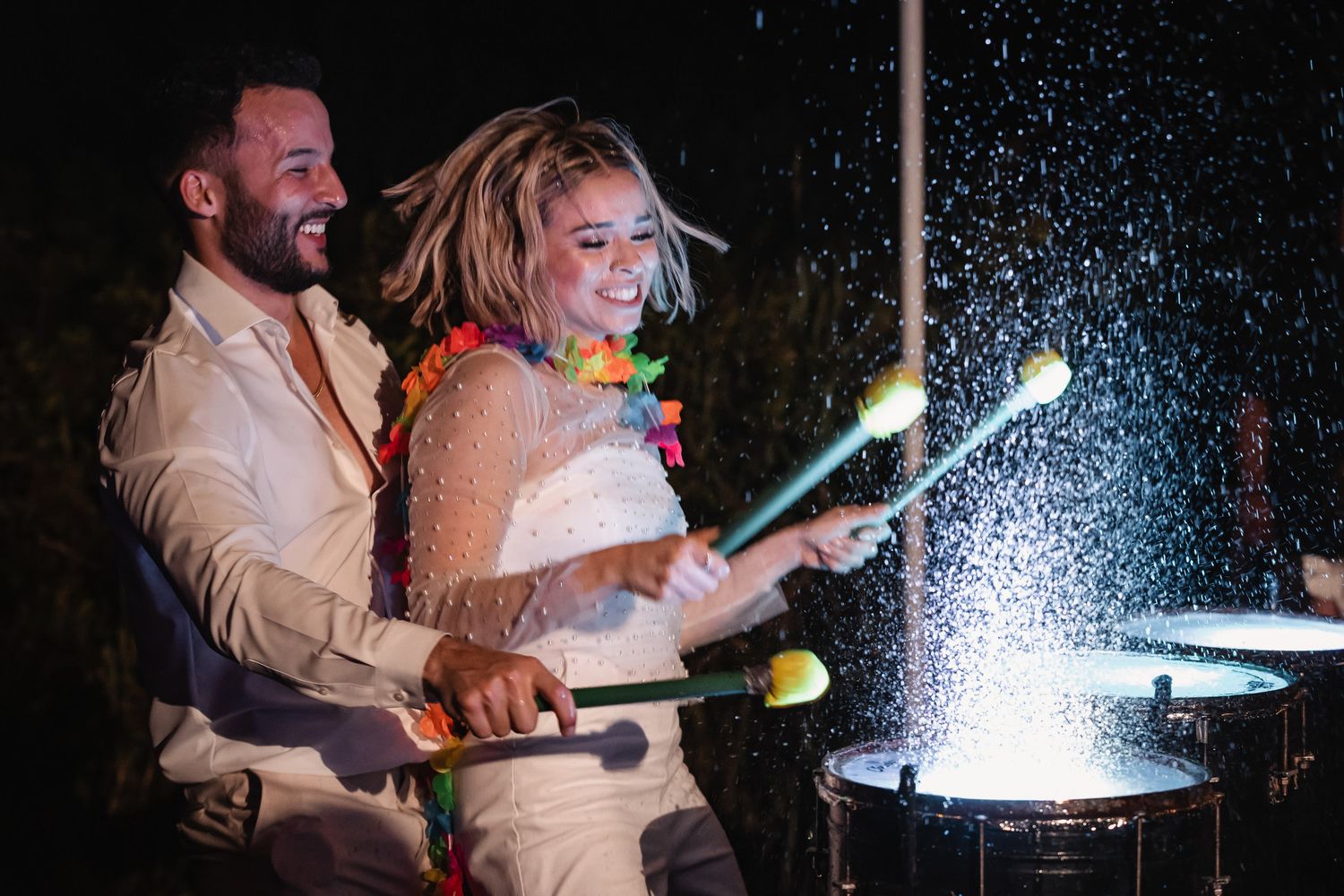 Bride and groom laughing while playing drums together at a lively outdoor wedding reception at night.