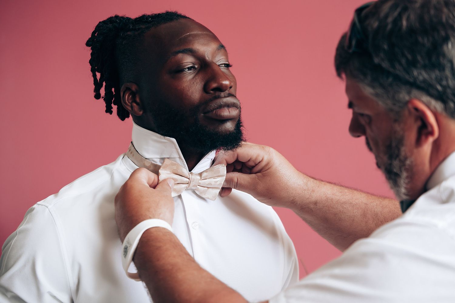 Someone adjusting a white bowtie on formal attire against a pink background.