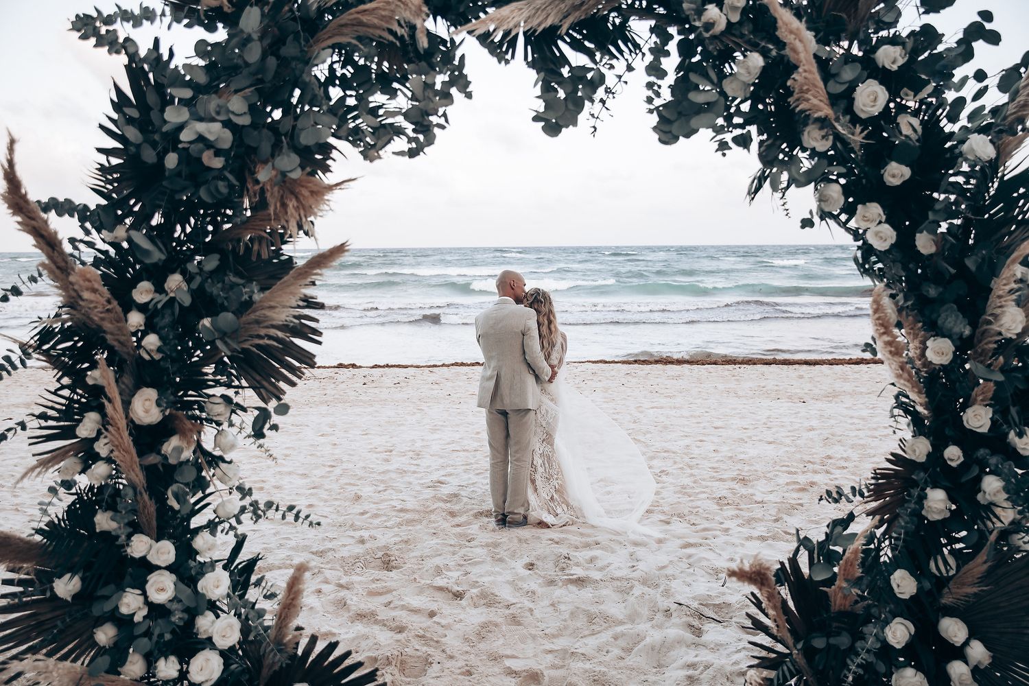 Wedding ceremony setup on a beach with decorative floral arch and ocean waves in the background.