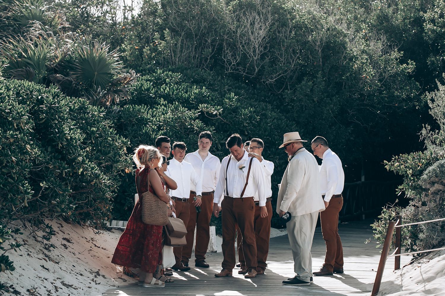 Wedding party members in light colored attire gather outdoors among trees.