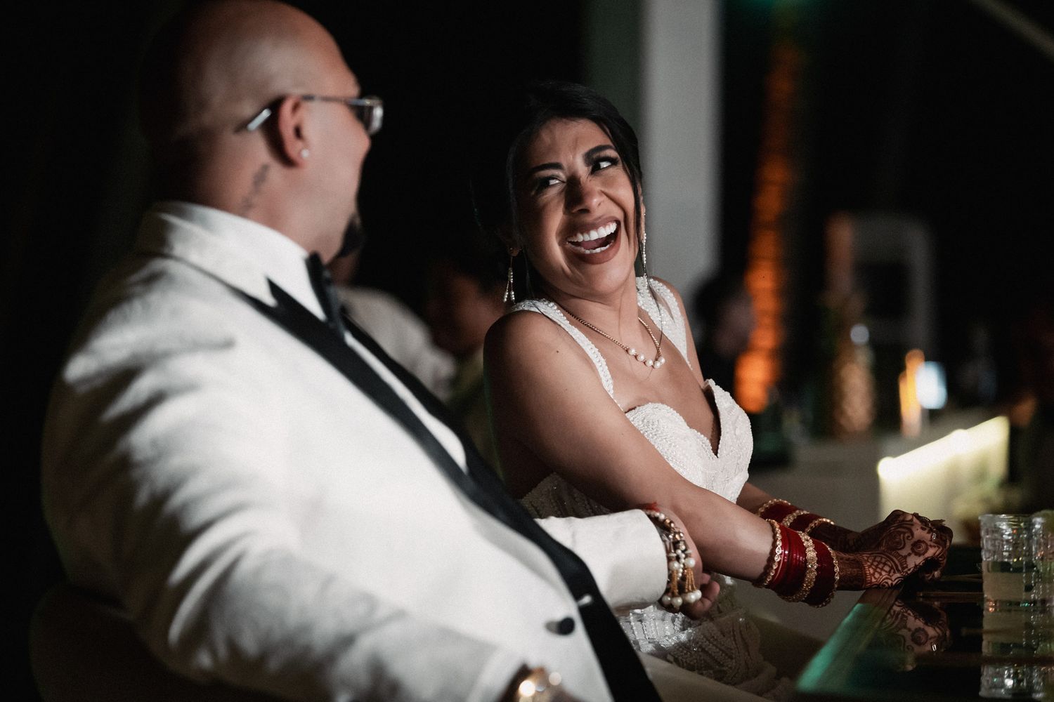 Bride in white gown laughing joyfully with groom in white suit at candlelit wedding reception table.