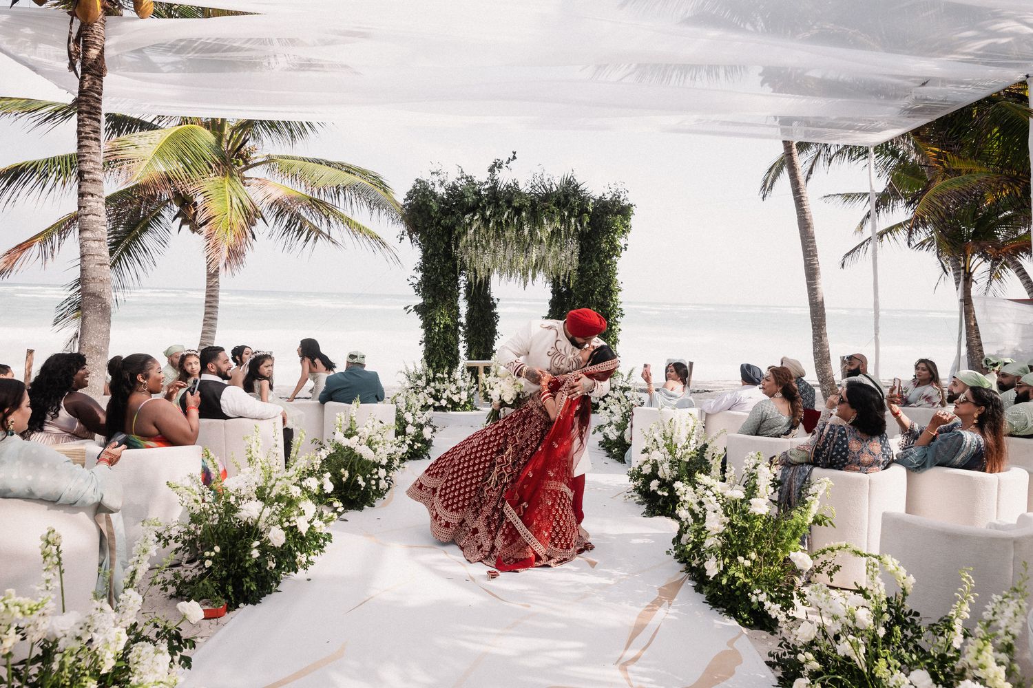 Bride in red lehenga walking down white aisle at outdoor beach wedding ceremony.