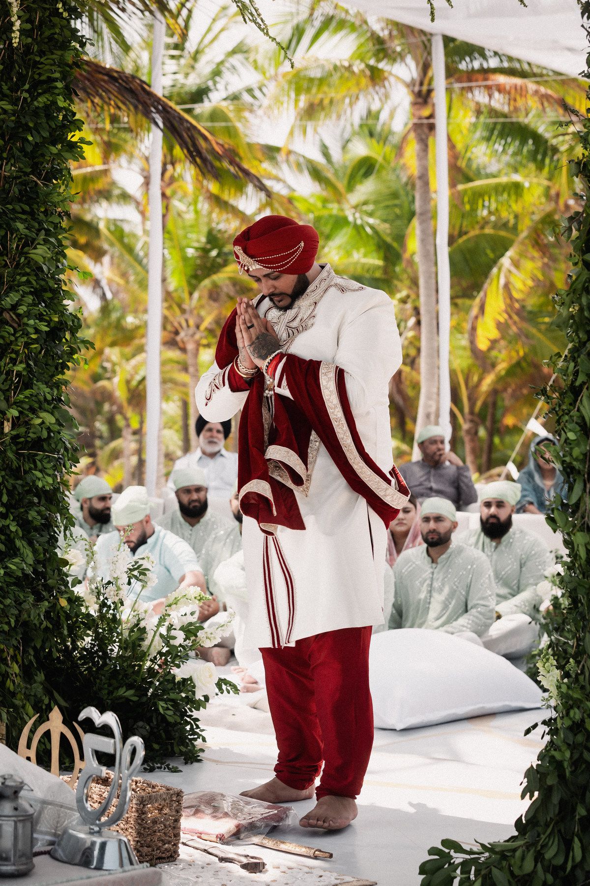 Sikh groom in white sherwani and red turban prays with folded hands at outdoor beach wedding ceremony.