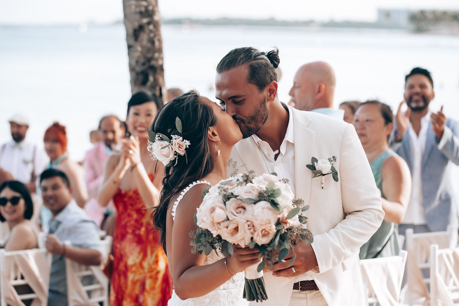 A tender moment between newlyweds during a beach wedding ceremony with guests watching.
