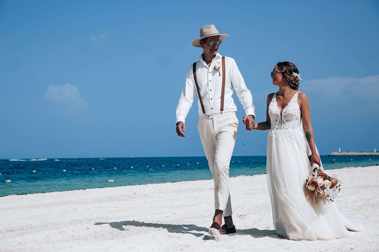 A couple in white outfits walking along a pristine beach with turquoise waters.