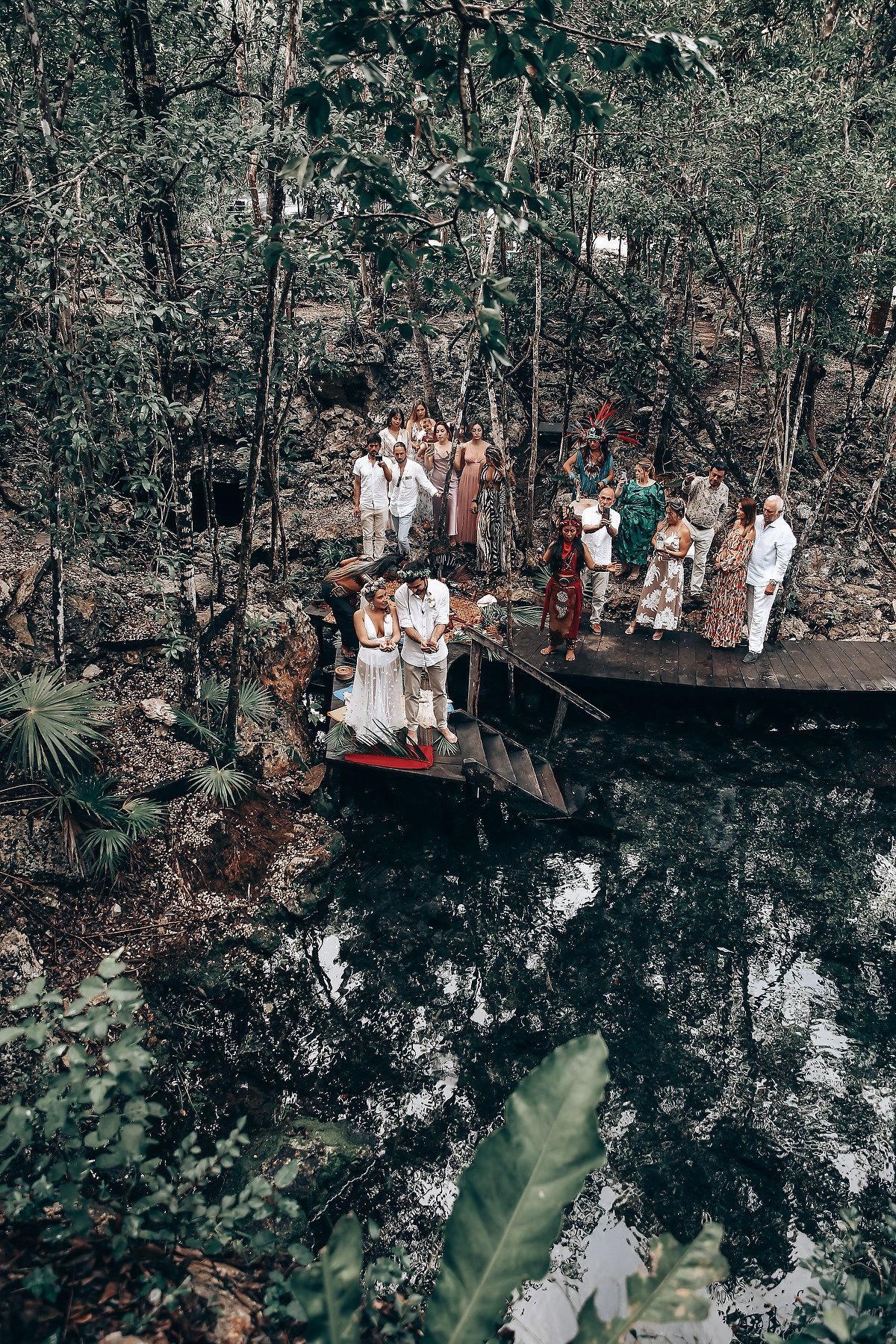 A group gathers on a wooden platform overlooking a dark cenote surrounded by lush jungle vegetation.