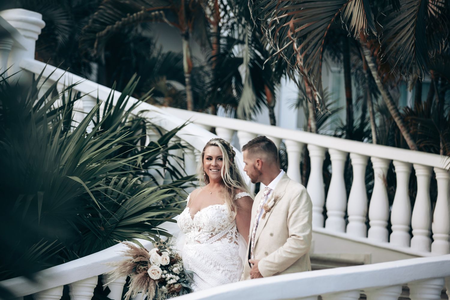 A smiling bride and groom share a romantic moment on a white balcony surrounded by lush tropical greenery.