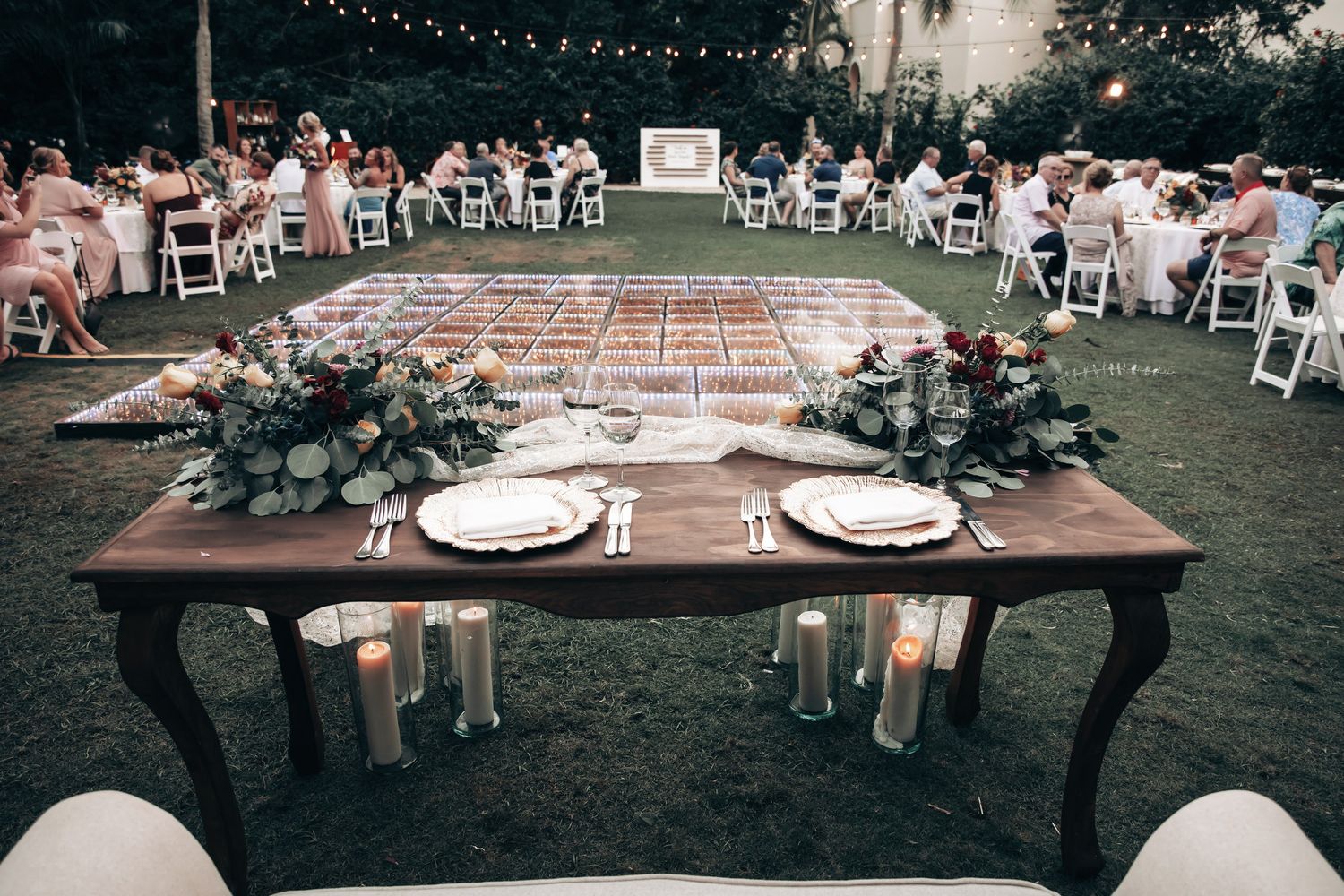 An elegant outdoor wedding reception with a sweetheart table, floral centerpieces, candles, and string lights overhead.