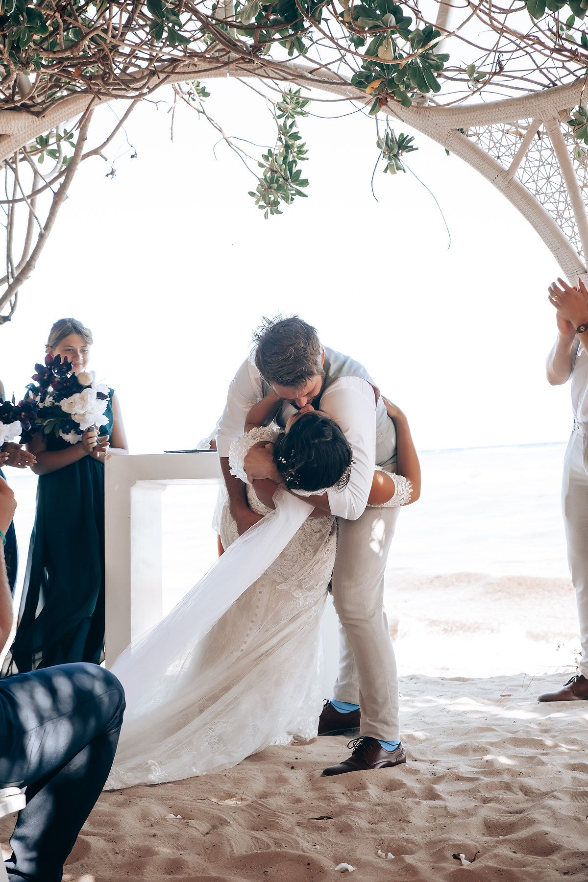 Wedding ceremony on sandy beach under wooden arch with flowing white fabric and greenery.