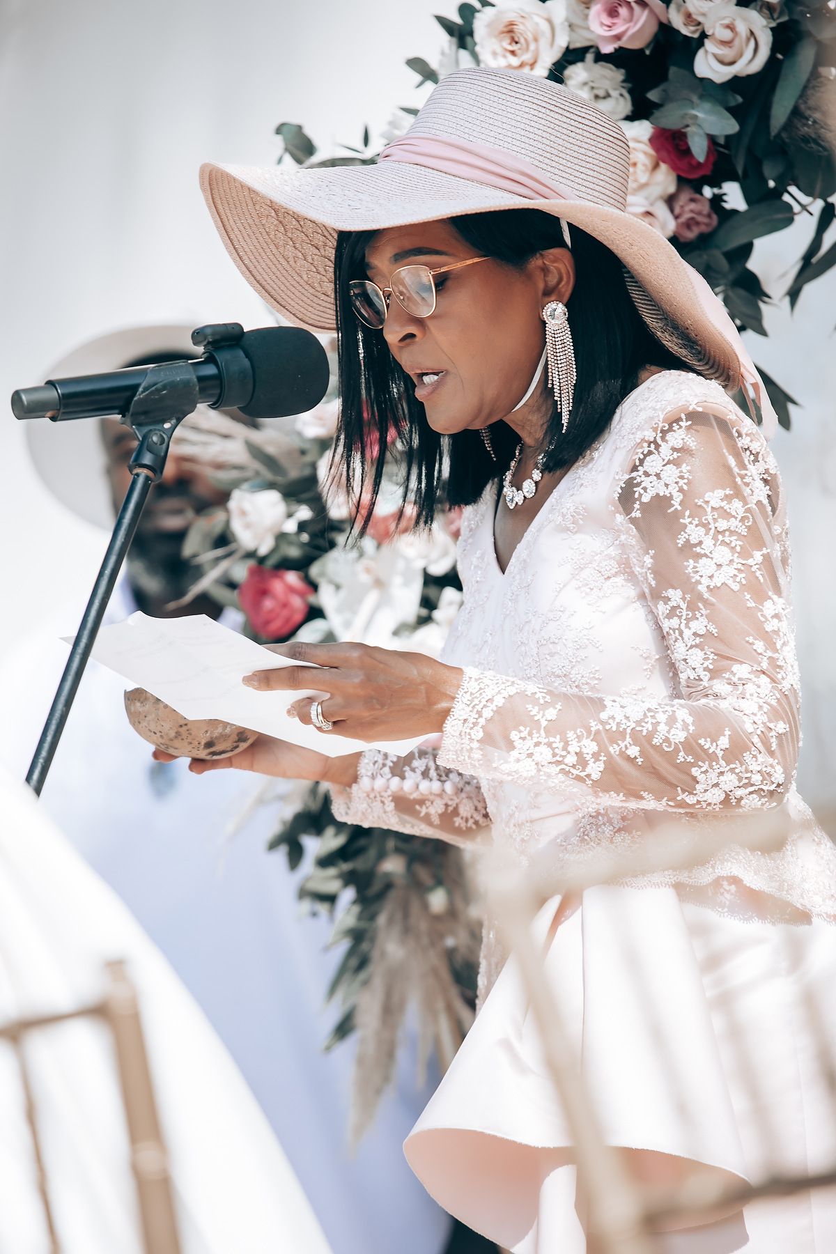 Elegant figure in white lace dress and wide-brimmed hat with floral decorations.