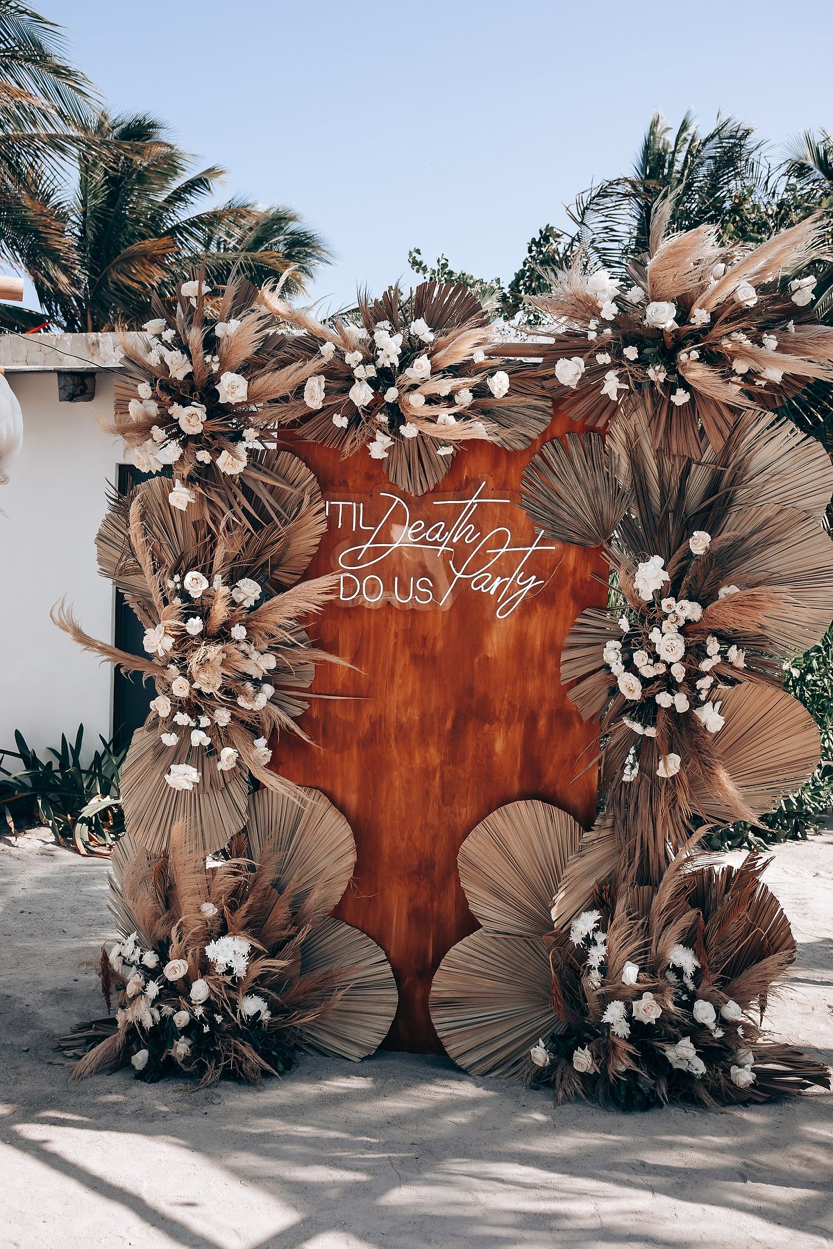Tropical wedding backdrop with dried palm fronds and white flowers arranged in a curved wooden wall display.