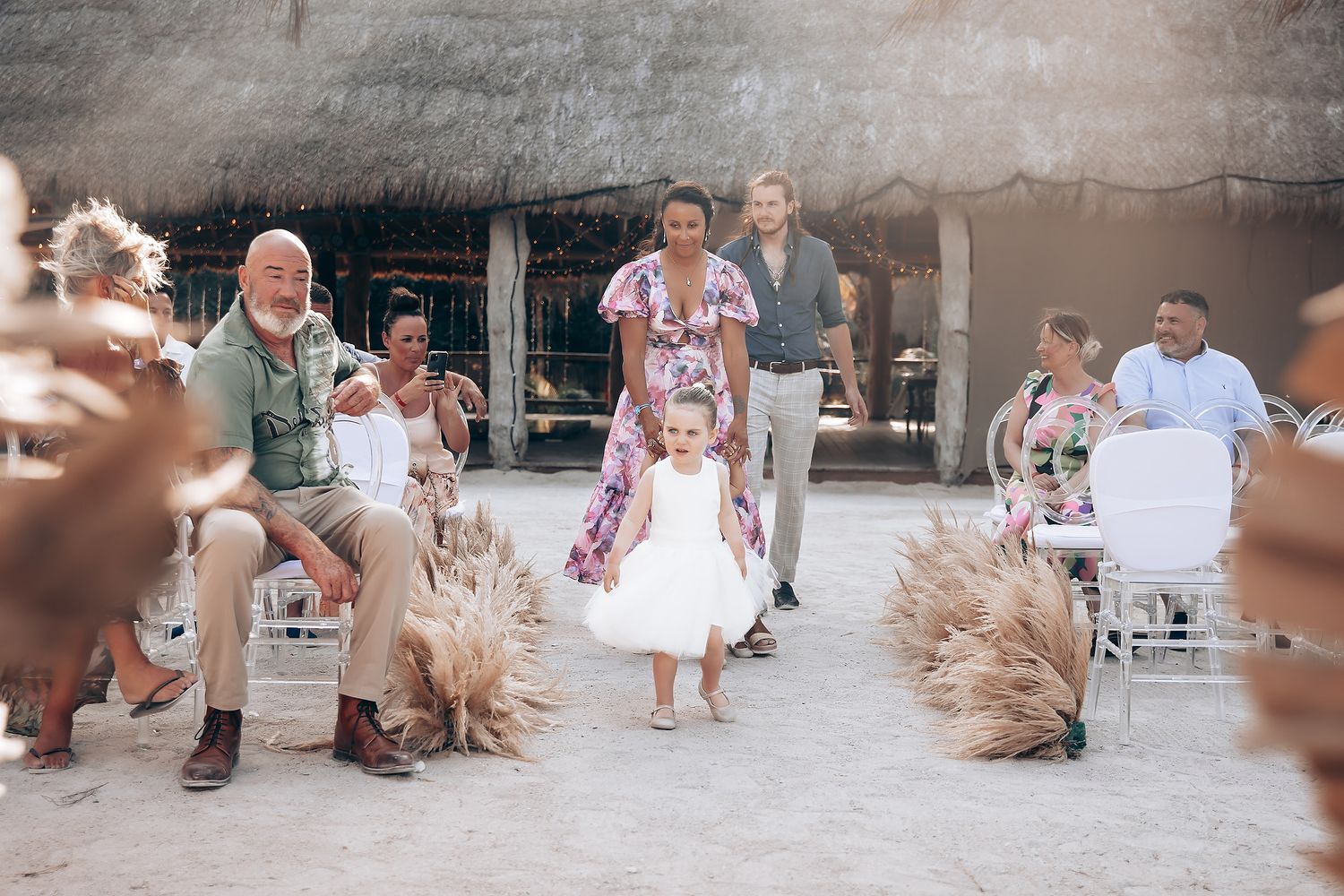 A wedding celebration with guests seated in white chairs surrounded by dried pampas grass decorations in a rustic venue.