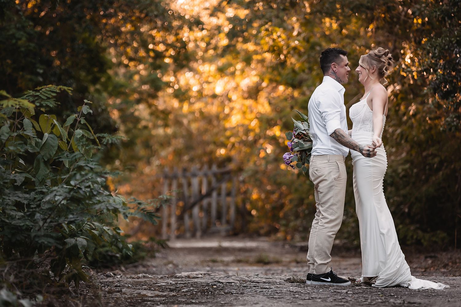 Bride and groom share a romantic moment on a sunlit forest path, bride holding purple bouquet in white gown.