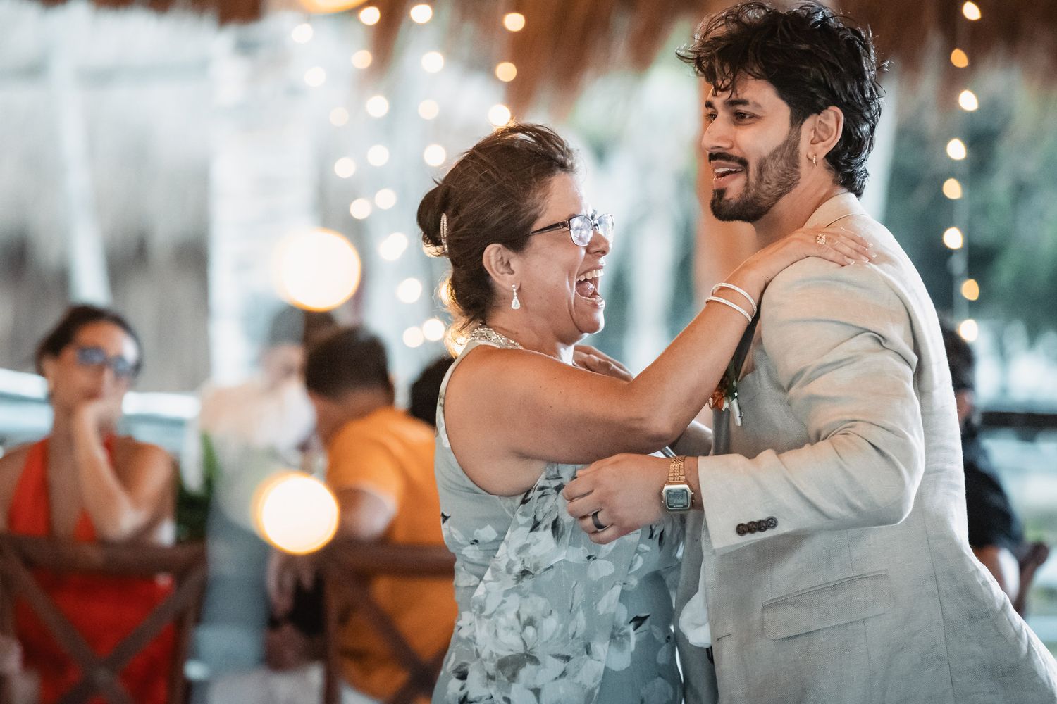Groom dances joyfully with older woman during outdoor beach wedding reception celebration.
