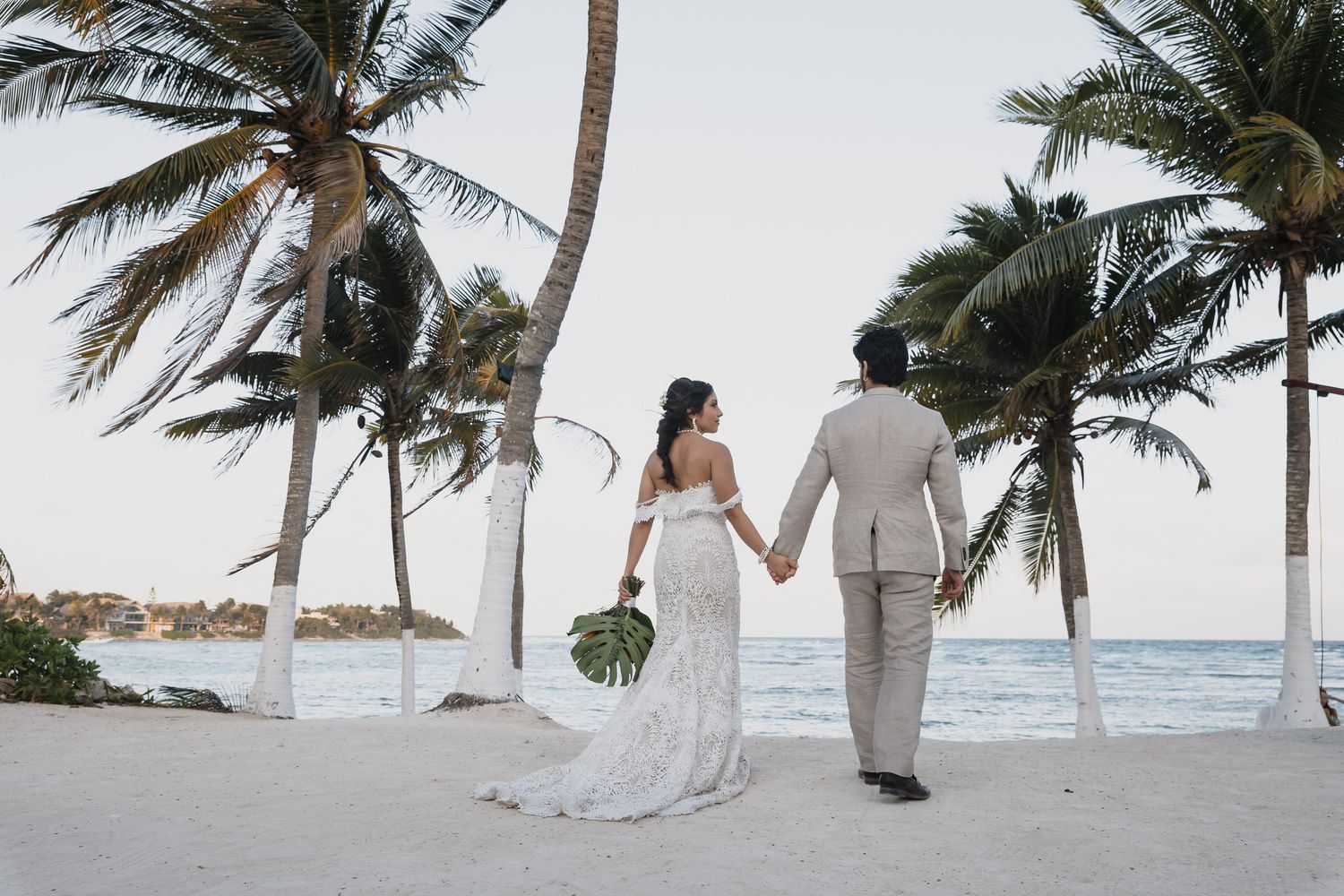 Bride and groom walk hand-in-hand on tropical beach with palm trees at sunset.