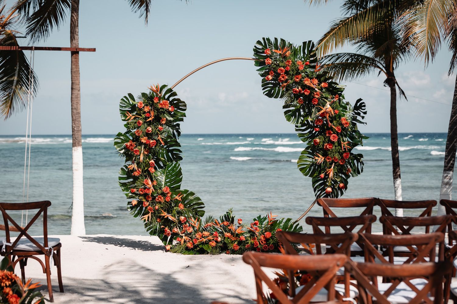 Tropical beach wedding ceremony setup with floral hoop arch adorned with green leaves and orange flowers, ocean backdrop.
