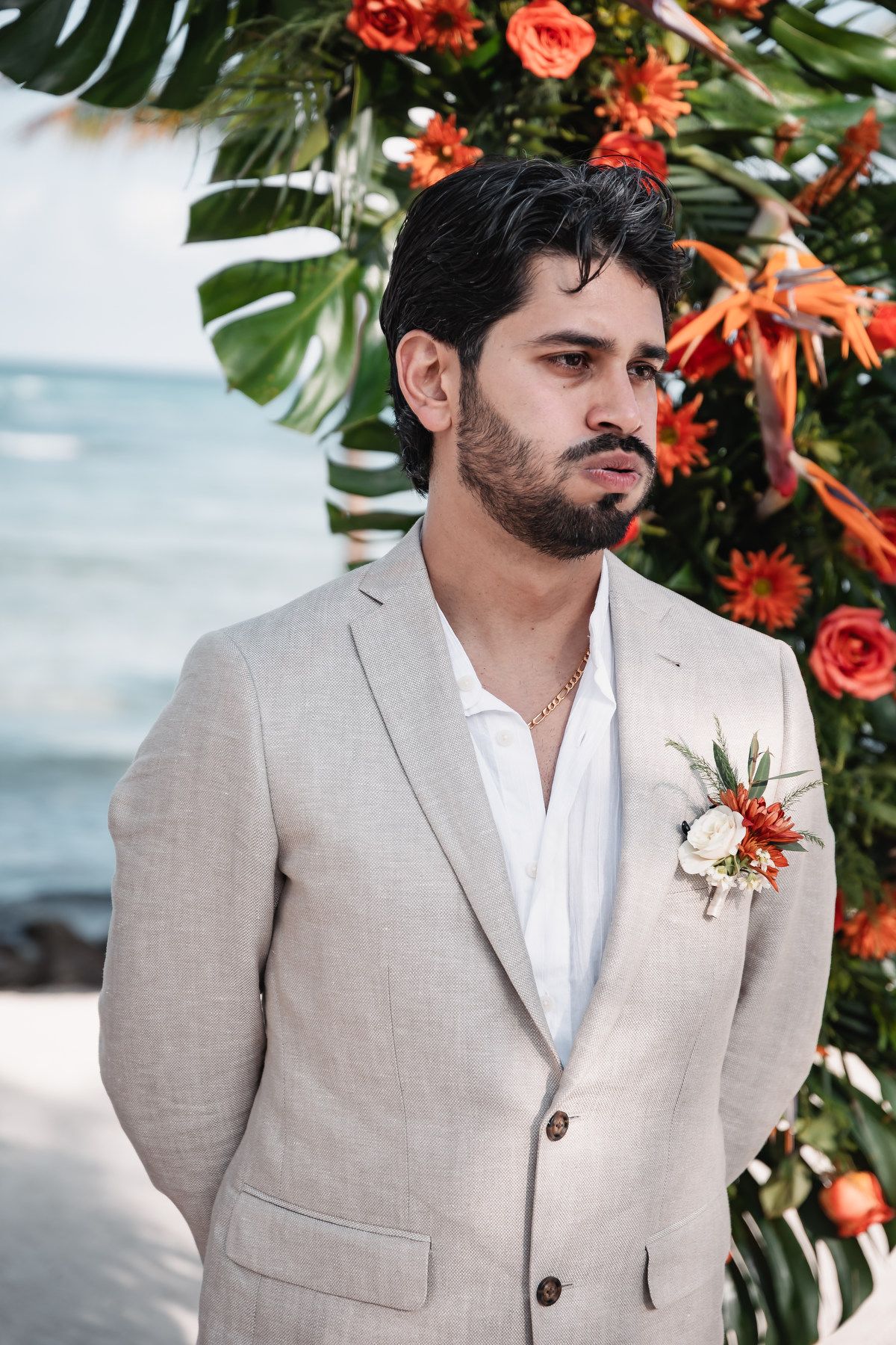 Handsome groom in linen suit with tropical boutonniere standing before colorful floral arch by the ocean.