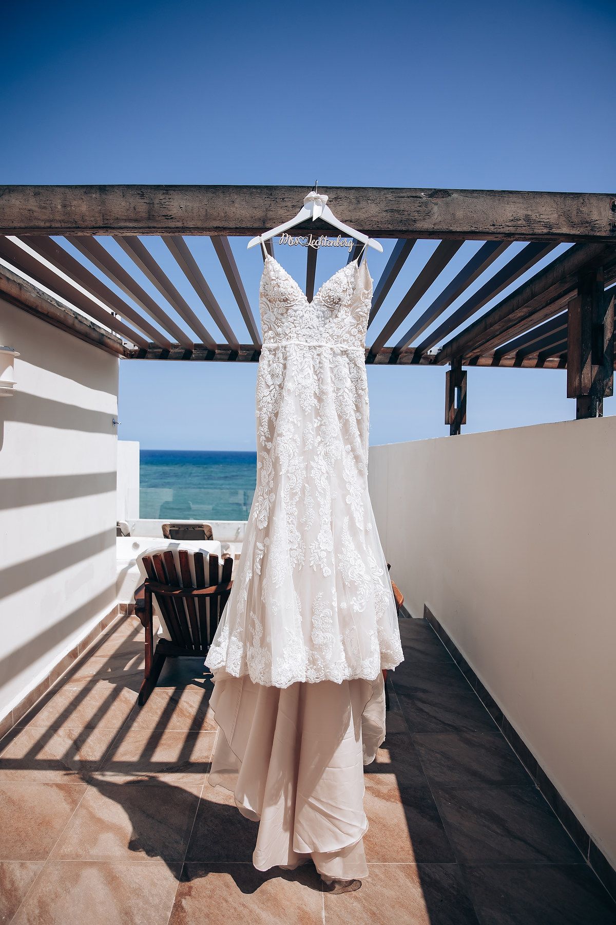 A white lace wedding dress hangs elegantly against a beachfront pergola with ocean views in the background.