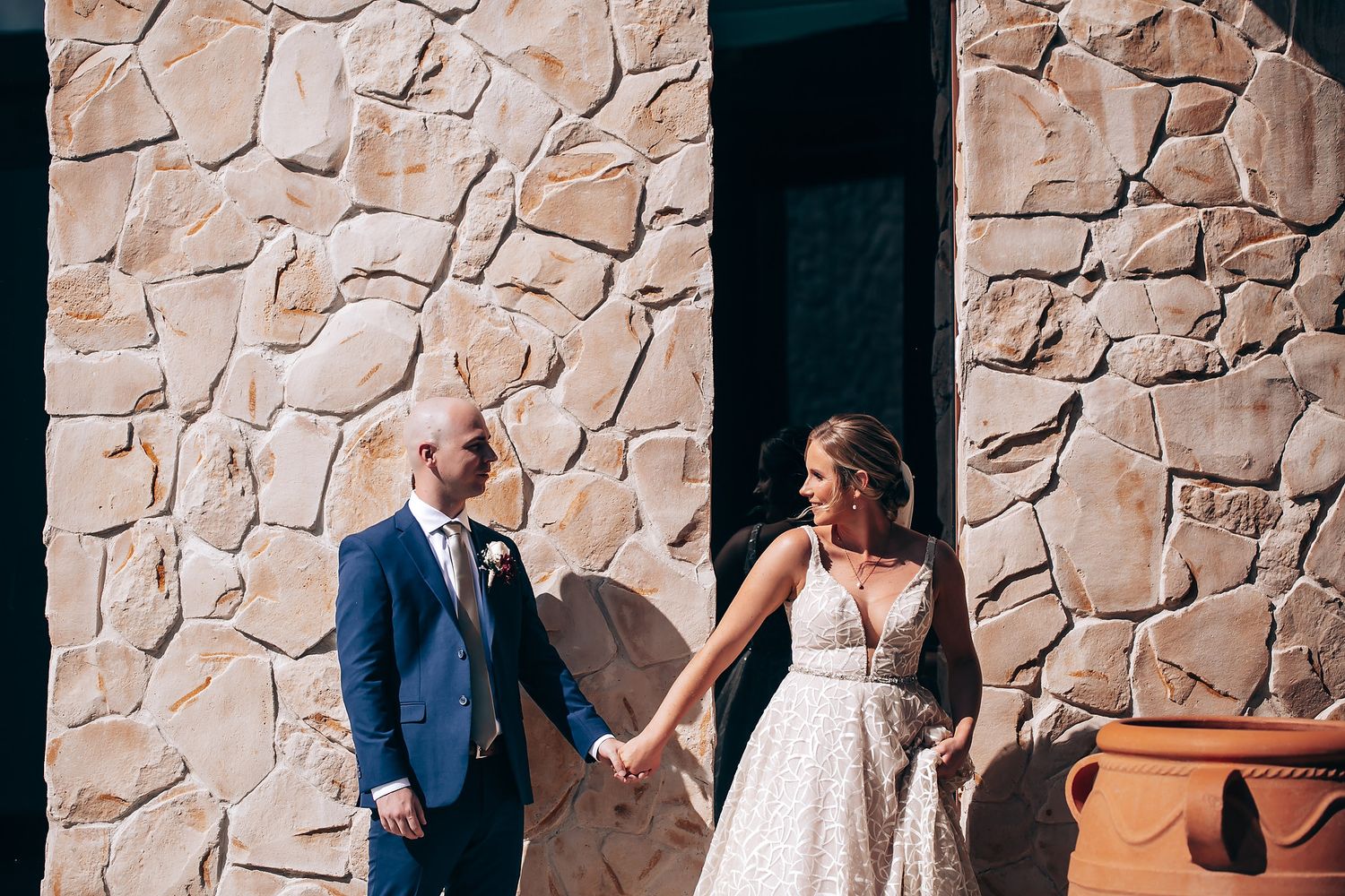 A bride and groom hold hands while standing in front of a stone wall on their wedding day.