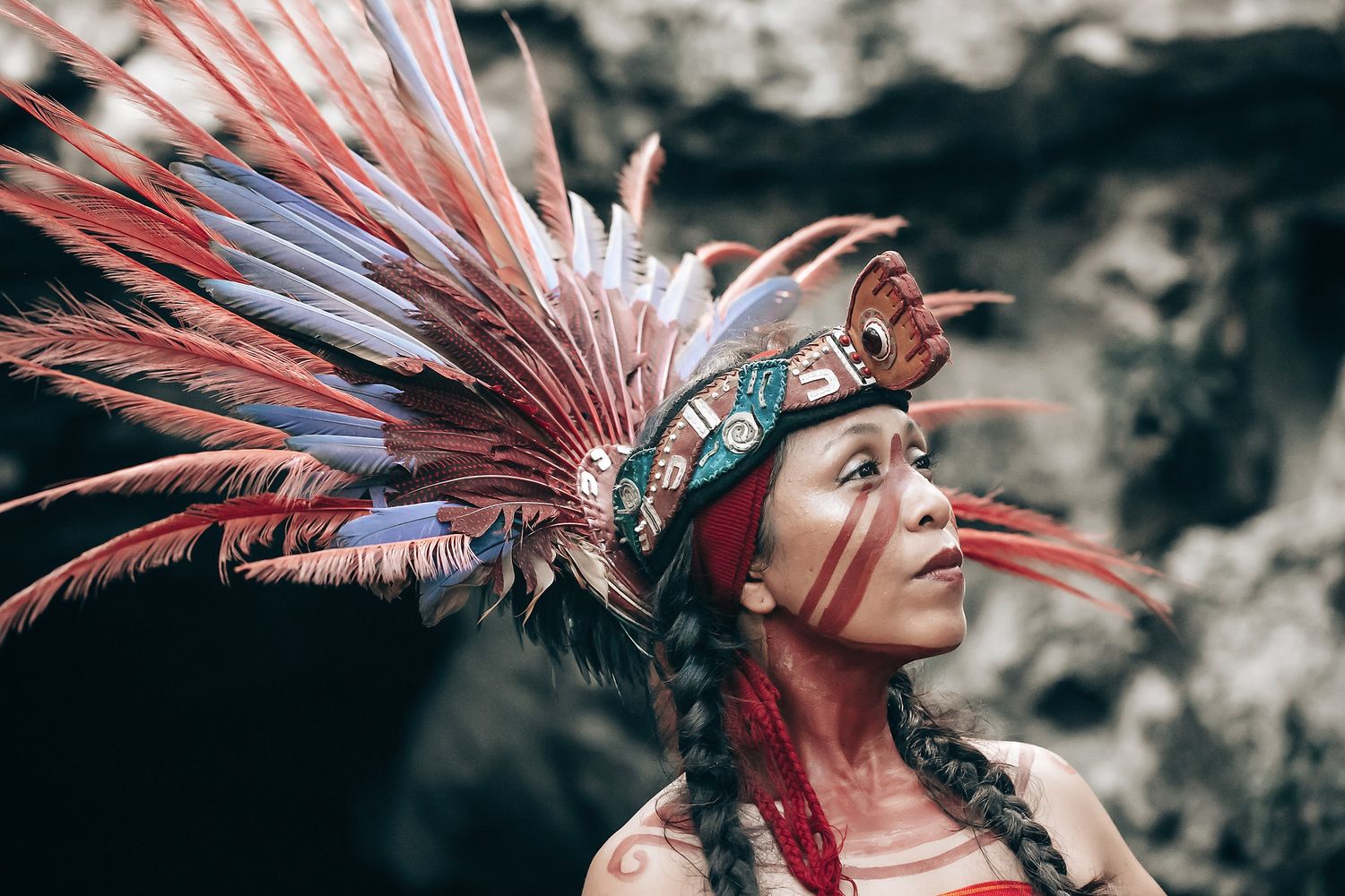A woman wearing a vibrant, feathered headdress stands in a rugged, natural environment.