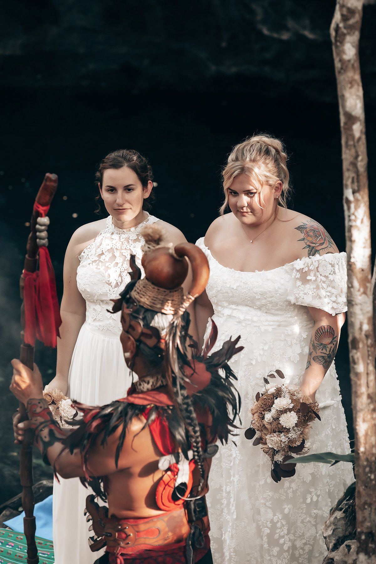 Two brides wearing white lace wedding dresses and holding bouquets stand in a lush, forested setting.