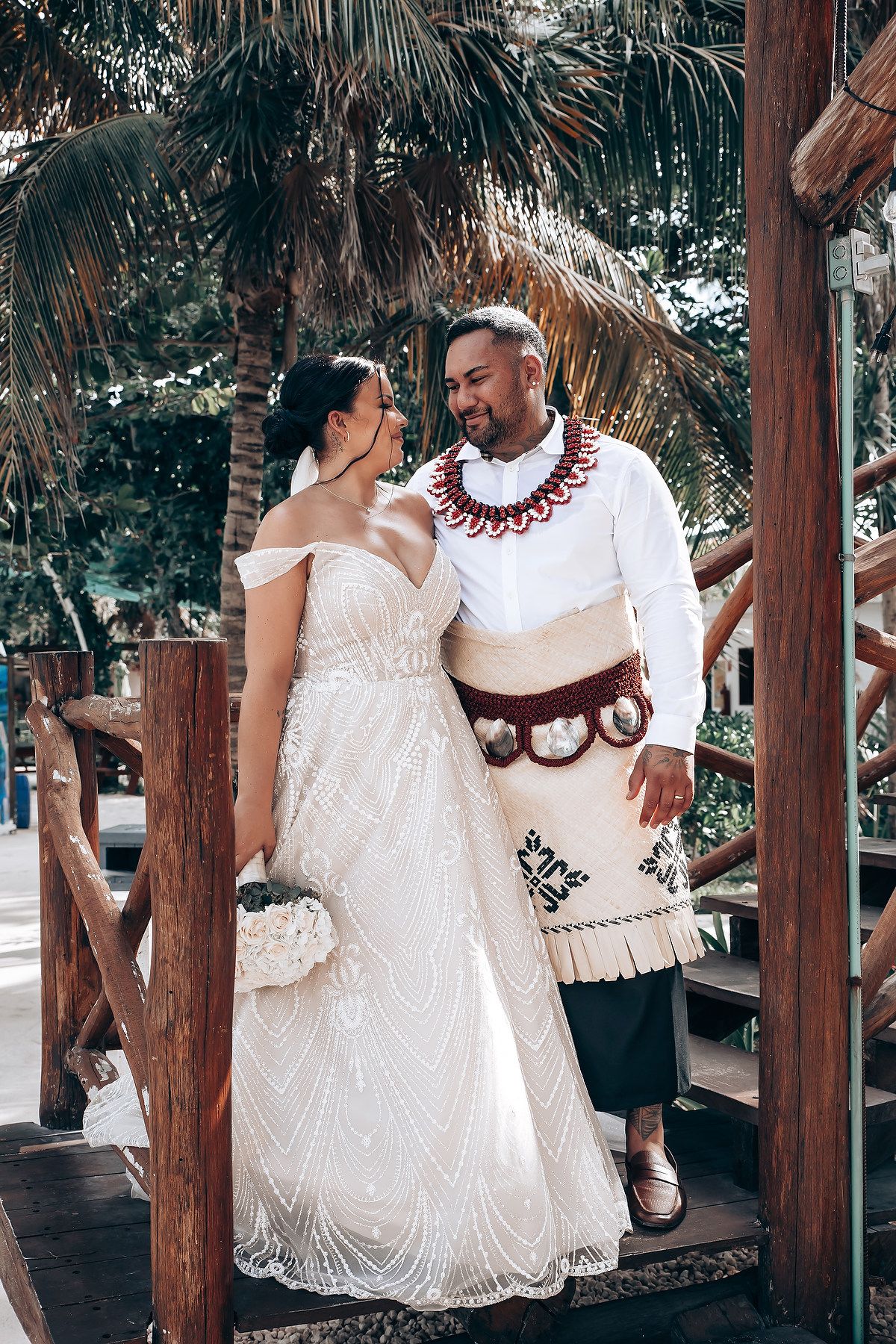 Couple in wedding attire sharing a moment under a rustic wooden pergola with palm trees.