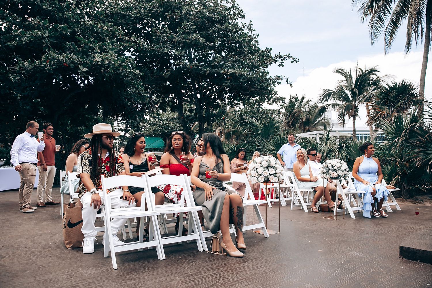 Wedding guests seated on white chairs at an outdoor tropical venue with palm trees in the background.