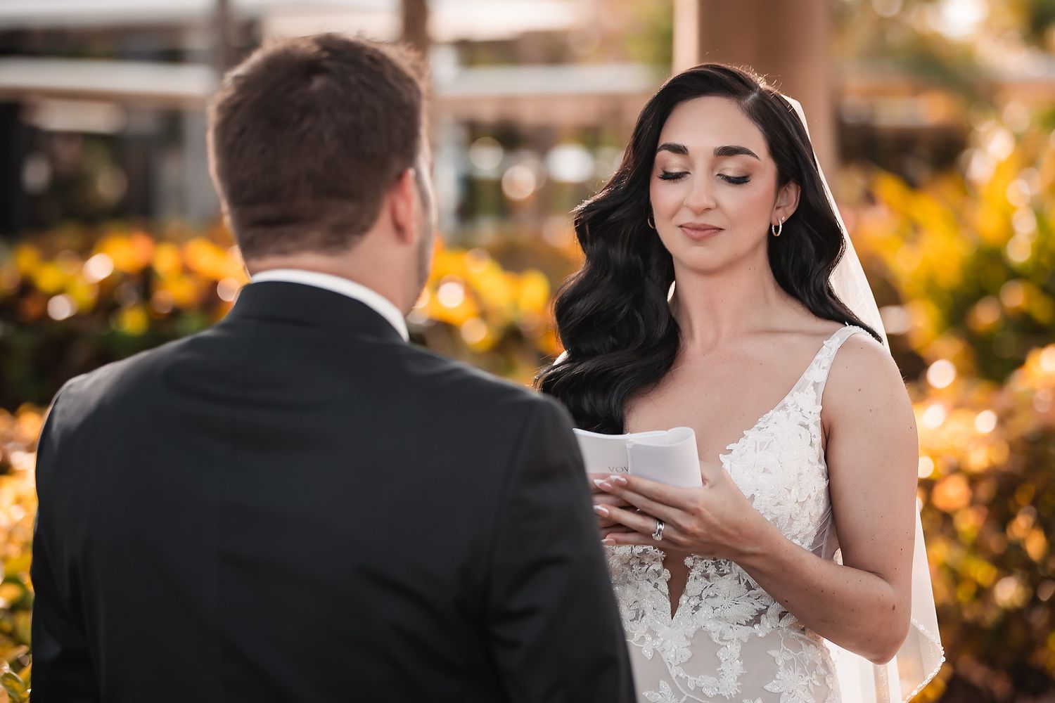 Bride in lace wedding dress reads vows to groom outdoors, with golden floral backdrop during wedding ceremony.